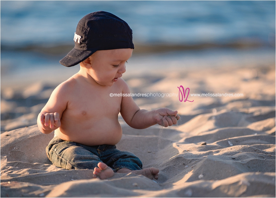 baby wearing baseball cap playing on the beach in the sand for the first time